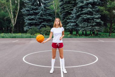 Güzel genç kız beyaz tişört, şort ve spor ayakkabısı, giyinmiş bir basketbol sahası üzerinde bir top ile çalış.