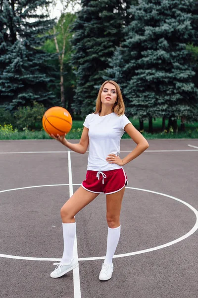 Hermosa joven vestida con camiseta blanca, pantalones cortos y zapatillas de deporte, juega con una pelota en una cancha de baloncesto . —  Fotos de Stock