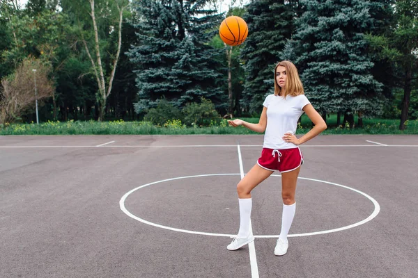 Belle jeune fille vêtue de t-shirt blanc, short et baskets, joue avec une balle sur un terrain de basket . — Photo
