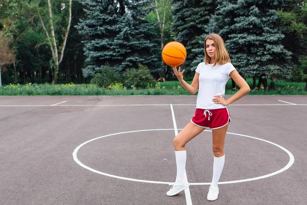 Smuk ung pige klædt i hvid t-shirt, shorts og sneakers, spiller med en bold på en basketballbane . - Stock-foto