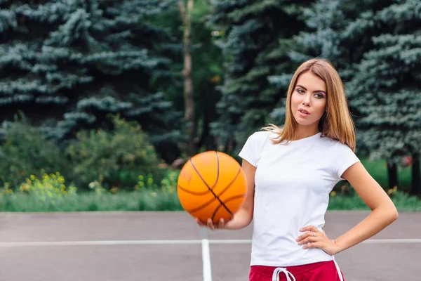 Krásná mladá dívka oblečená v bílé tričko, kraťasy a tenisky, hraje s míčem na basketbalové hřiště. — Stock fotografie