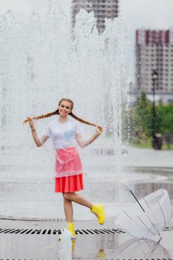 Genç güzel kız sarı çizmeli iki örgüler ve şeffaf şemsiye standları Çeşme yakınındaki.