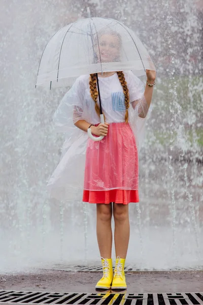 Young pretty girl with two braids in yellow boots and with transparent umbrella stands near fountain. — Stock Photo, Image