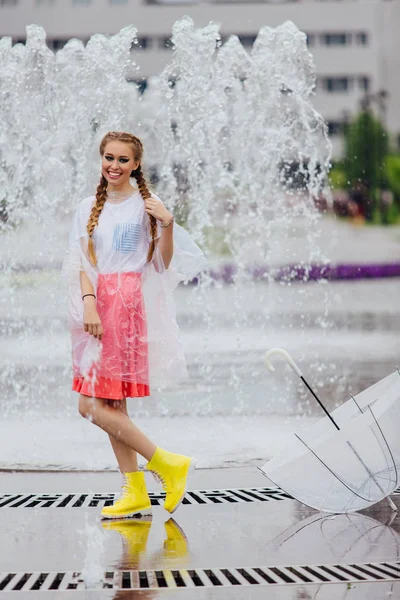 Joven chica bonita con dos trenzas en botas amarillas y con paraguas transparente se levanta cerca de la fuente . — Foto de Stock