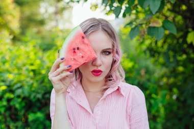Bir dilim karpuz yüzünü önünde tutan pembe saçlı güzel genç kadın
