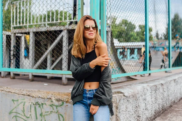 Retrato de moda de una joven de moda con gafas de sol, jeans con pasillos y chaqueta de bombardero en la ciudad —  Fotos de Stock