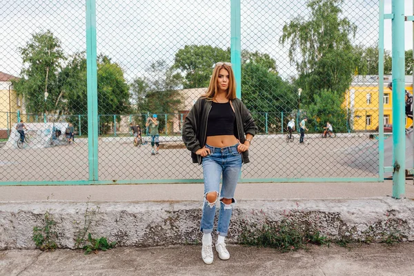 Potret busana wanita muda trendi mengenakan kacamata hitam, dan jaket pembom duduk di sebelah rabitz di kota — Stok Foto