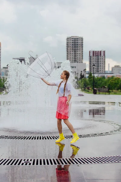 Junges hübsches Mädchen mit zwei Zöpfen in gelben Stiefeln wirft und fängt transparenten Regenschirm in der Nähe des Brunnens. — Stockfoto