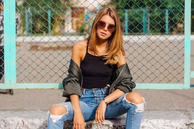 Güneş gözlüğü ve sıva şehrin yanında oturan bombardıman ceket giyen trendy genç kadın moda portre