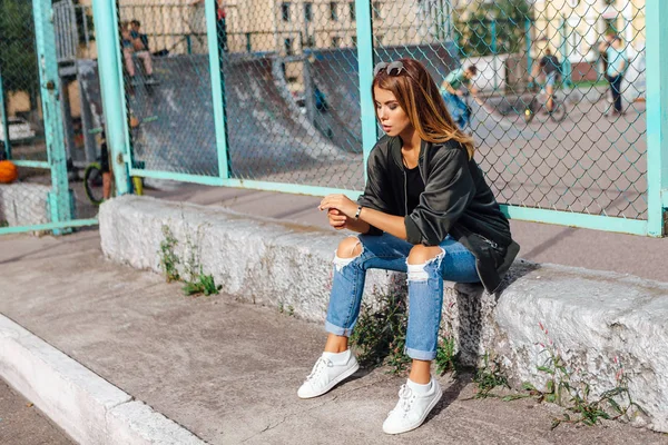 Fashion portrait of trendy young woman wearing sunglasses, and bomber jacket sitting next to rabitz in the city — Stock Photo, Image