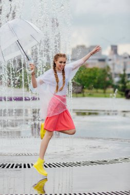 Genç güzel kız sarı çizmeli iki örgüler ve şeffaf şemsiye standları Çeşme yakınındaki.