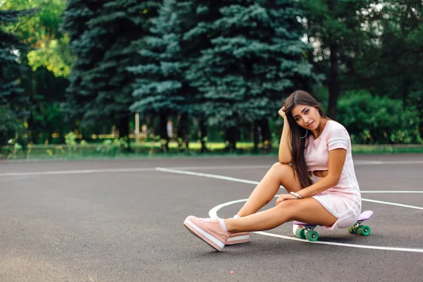 Portrait of a smiling charming brunette female sitting on her skateboard on a basketball court. — Stock Photo, Image