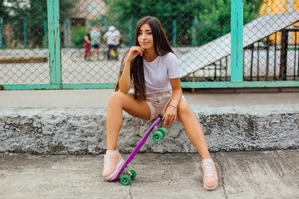 Zomer levensstijl foto van trendy mooie jonge meisje zit naast de skateboard coart met haar kunststof skateboard. — Stockfoto