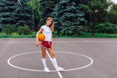 Güzel genç kız beyaz tişört, şort ve spor ayakkabısı, giyinmiş bir basketbol sahası üzerinde bir top ile çalış.