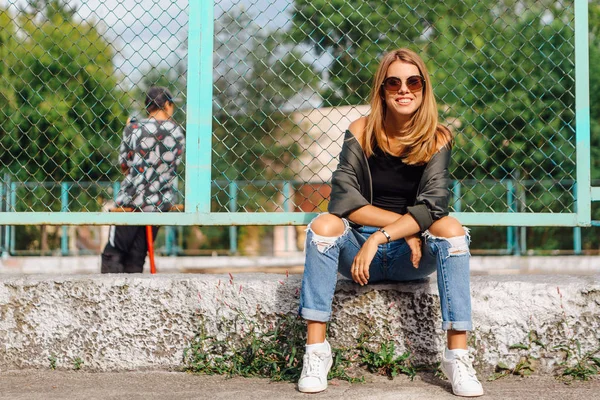 Fashion portrait of trendy young woman wearing sunglasses, and bomber jacket sitting next to rabitz in the city — Stock Photo, Image