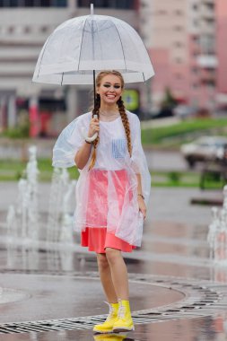 Genç güzel kız sarı çizmeli iki örgüler ve şeffaf şemsiye standları Çeşme yakınındaki.