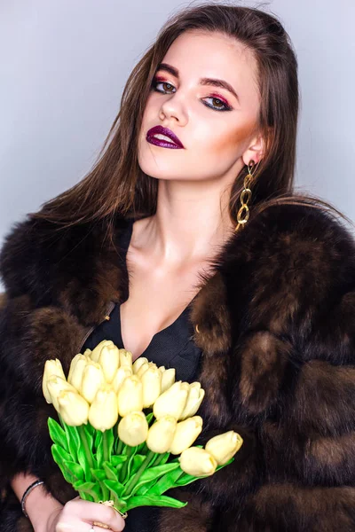 Retrato de belleza de una mujer con maquillaje de colores brillantes vestida con un abrigo de piel que sostiene el ramo de tulipanes amarillos — Foto de Stock