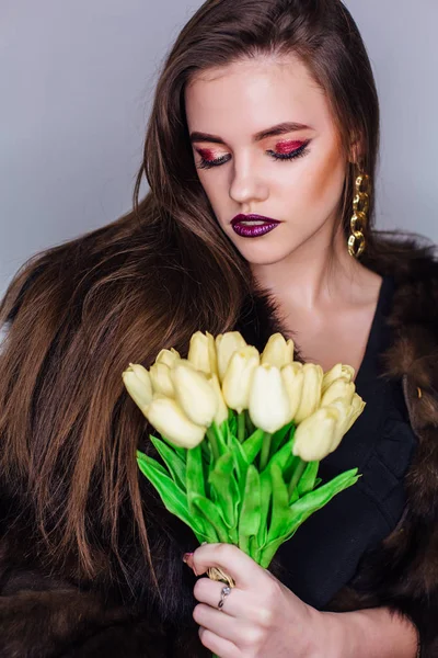 Retrato de belleza de una mujer con maquillaje de colores brillantes vestida con un abrigo de piel que sostiene el ramo de tulipanes amarillos — Foto de Stock
