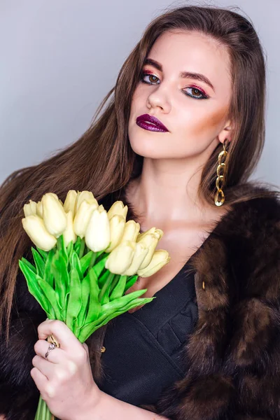 Ritratto di bellezza di una donna con brillante trucco colorato vestito con una pelliccia che tiene bouquet di tulipani gialli — Foto Stock