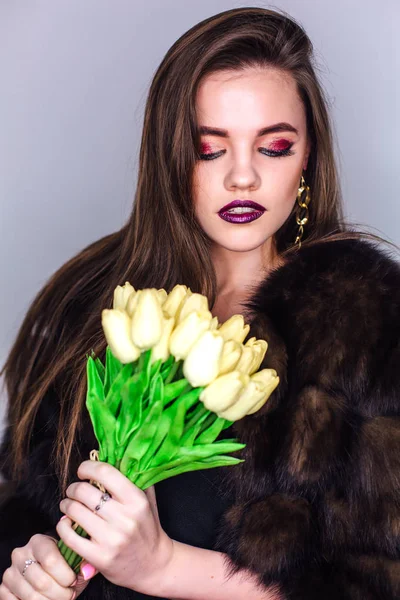 Retrato de belleza de una mujer con maquillaje de colores brillantes vestida con un abrigo de piel que sostiene el ramo de tulipanes amarillos — Foto de Stock