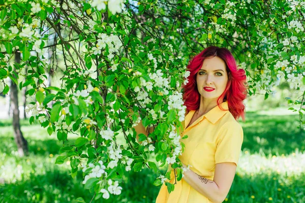 Όμορφη και των νέων γυναίκας με ζωηρά κόκκινα χείλια μαλλιά και ref στέκεται δίπλα σε ένα ανθισμένο δέντρο της apple σε ένα κίτρινο φόρεμα. — Φωτογραφία Αρχείου