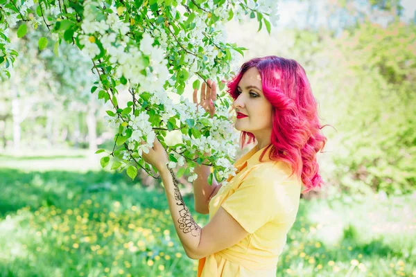 Vacker och ung kvinna med röda hår och ref läppar står bredvid en blommande äppelträd i en gul klänning. — Stockfoto