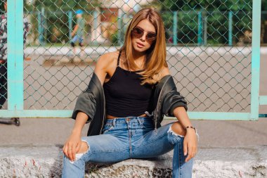 Güneş gözlüğü ve sıva şehrin yanında oturan bombardıman ceket giyen trendy genç kadın moda portre