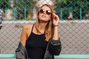 Güneş gözlüğü ve sıva şehrin yanında oturan bombardıman ceket giyen trendy genç kadın moda portre