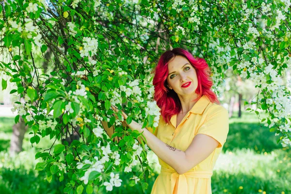 Mooie en jonge vrouw met heldere rode haren en ref lippen permanent naast een bloeiende appelboom in een gele jurk. — Stockfoto