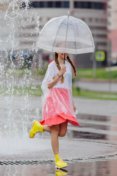 Jonge mooie meisje met twee vlechten in gele laarzen en met transparante paraplu stands in de buurt van fontein. — Stockfoto