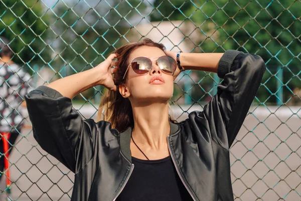 Retrato de moda de jovem na moda usando óculos de sol, e jaqueta de bombardeiro sentado ao lado de rabitz na cidade — Fotografia de Stock