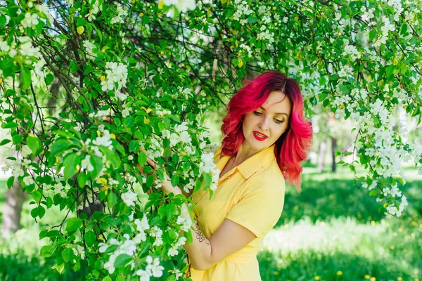 Красива і молода жінка з яскраво-червоним волоссям і реф губами, що стоять поруч з квітучою яблунею в жовтій сукні . — стокове фото
