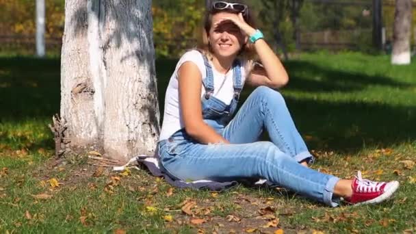 Mujer sonriente en mono jaens y zapatillas rojas sentadas debajo de un árbol en el parque de otoño — Vídeos de Stock