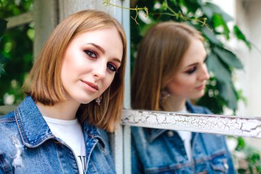 Denim ceket ve güzel kulak halkaları ayna penceresi yakınında duran moda kız, onun yansıması waching.