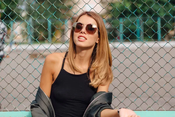 Modeporträt einer trendigen jungen Frau mit Sonnenbrille und Bomberjacke, die neben rabitz in der Stadt sitzt — Stockfoto