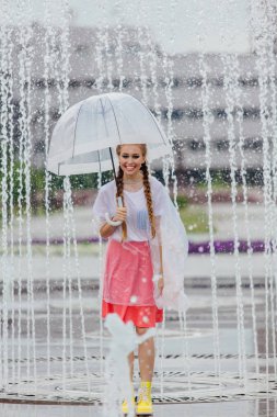 Genç güzel kız sarı çizmeli iki örgüler ve şeffaf şemsiye standları Çeşme yakınındaki.