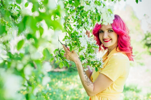 Mulher bonita e jovem com cabelos vermelhos brilhantes e lábios rep pé ao lado de uma macieira florescente em um vestido amarelo . — Fotografia de Stock