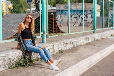 Güneş gözlüğü ve sıva şehrin yanında oturan bombardıman ceket giyen trendy genç kadın moda portre