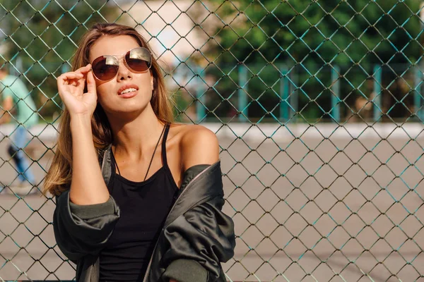 Güneş gözlüğü ve sıva şehrin yanında oturan bombardıman ceket giyen trendy genç kadın moda portre — Stok fotoğraf