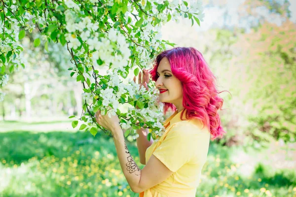 Schöne junge Frau mit leuchtend roten Haaren und ref Lippen, die neben einem blühenden Apfelbaum in einem gelben Kleid steht. — Stockfoto