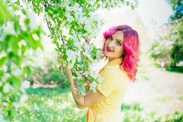 Piękna i młoda kobieta z jasne czerwone włosy i ref usta stojący obok kwitnących jabłoni w żółtej sukience. — Zdjęcie stockowe