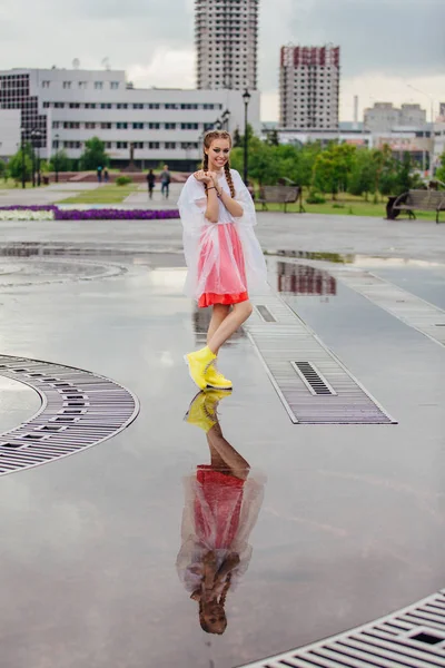 Menina bonita molhada nova com duas tranças em botas amarelas fica perto da fonte. Menina refletindo no chão molhado — Fotografia de Stock