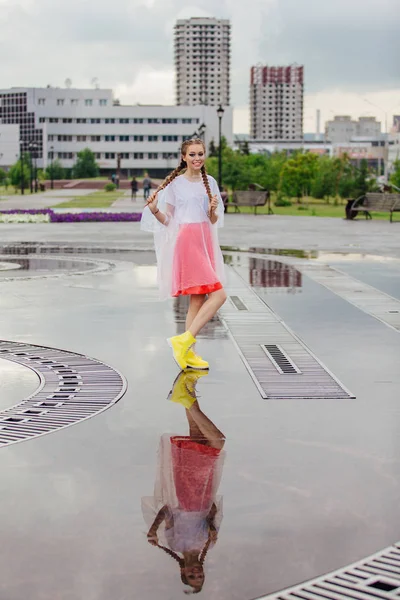 Gadis cantik basah muda dengan dua kepang sepatu bot kuning berdiri di dekat air mancur. Gadis muda mencerminkan di tanah basah — Stok Foto