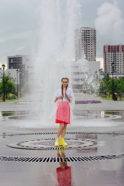 Joven chica bonita mojada con dos trenzas en botas amarillas y con paraguas transparente en el interior de la fuente . — Foto de Stock