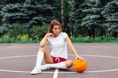 Beyaz tişört, şort ve spor ayakkabıları giymiş güzel genç kız, topu ile bir basketbol sahası oturur.