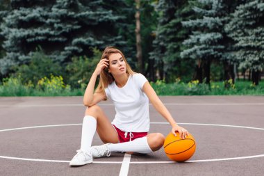Beyaz tişört, şort ve spor ayakkabıları giymiş güzel genç kız, topu ile bir basketbol sahası oturur.