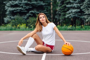 Beyaz tişört, şort ve spor ayakkabıları giymiş güzel genç kız, topu ile bir basketbol sahası oturur.