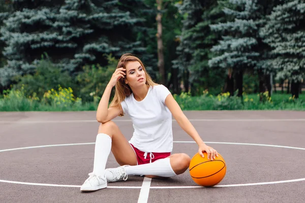 Gadis muda cantik berpakaian putih t-shirt, celana pendek dan sepatu olahraga, duduk di lapangan basket dengan bola . — Stok Foto