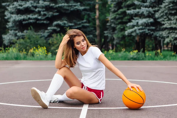 Schönes junges Mädchen in weißem T-Shirt, kurzer Hose und Turnschuhen, sitzt auf einem Basketballfeld mit Ball. — Stockfoto