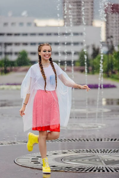 Joven chica bonita mojada con dos trenzas en botas amarillas y con paraguas transparente en el interior de la fuente . — Foto de Stock